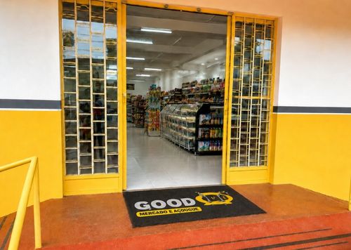 Fachada de mercado de bairro com portas de vidro decoradas e amarelas, capacho personalizado preto e amarelo com a logomarca Good Mercado e Açougue na entrada, piso de cerâmica vermelha, corredores com prateleiras ao fundo, fotografia realista de fachada comercial em dia claro