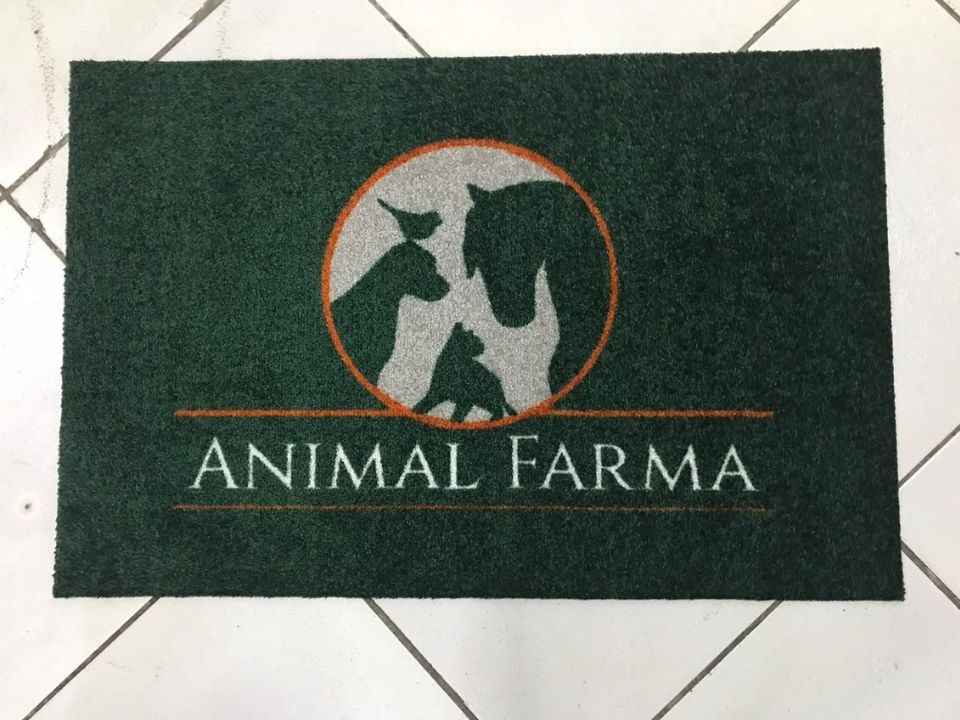 Capacho personalizado verde escuro com um círculo prateado contendo silhuetas de animais (cão, gato, pássaro e cavalo) em contorno laranja, e o texto Animal Farma em branco abaixo, sobre piso de cerâmica clara, fotografia realista de tapete para clínica veterinária