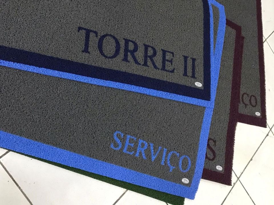 Dois tapetes personalizados cinza com bordas azuis e vinho, um com o texto Torre II Serviço em azul marinho e outro com Serviço em bordô, dispostos em diagonal sobre piso de cerâmica branca, fotografia realista de tapetes de identificação para condomínio
