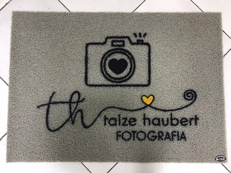 Capacho personalizado cinza claro com ilustração de câmera fotográfica com coração e a assinatura Taize Haubert Fotografia em preto e dourado, sobre piso de cerâmica clara, fotografia realista de tapete para estúdio criativo