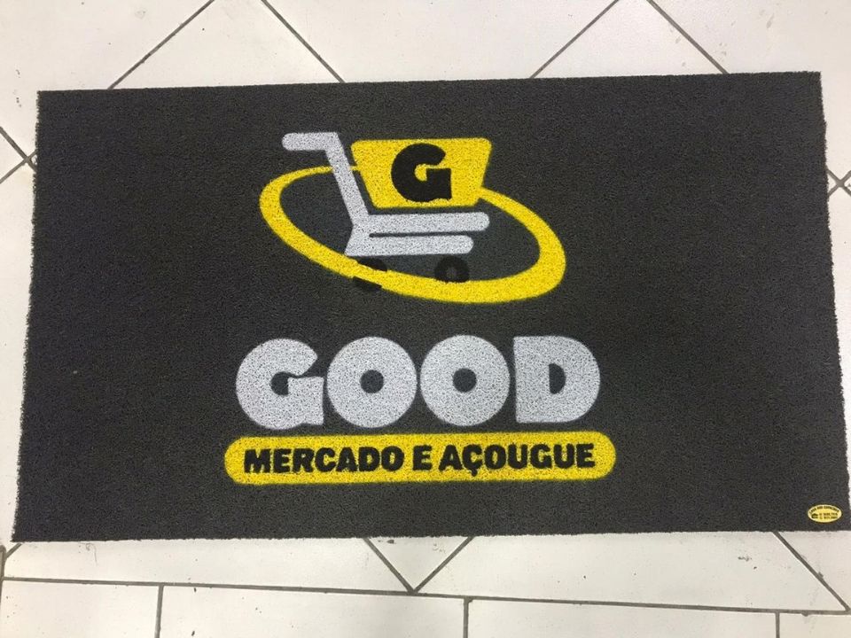 Capacho personalizado preto com a logomarca Good Mercado e Açougue em amarelo, branco e preto, textura antiderrapante de fibra densa, sobre piso de cerâmica clara, fotografia realista de tapete comercial resistente