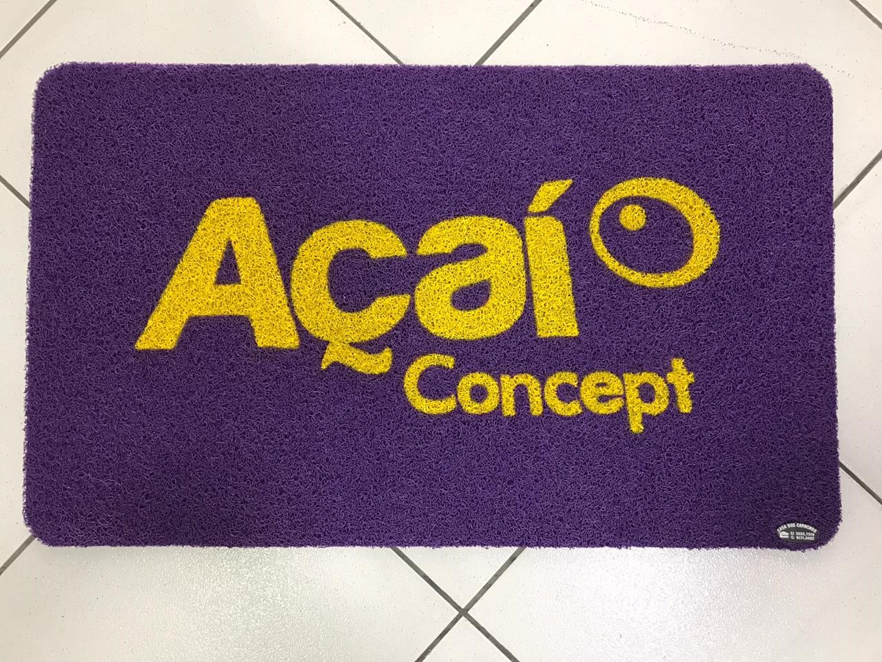 Capacho personalizado roxo com a logomarca Açaí Concept em letras amarelas, textura de fibra colorida, sobre piso de cerâmica clara, fotografia realista de tapete comercial vibrante