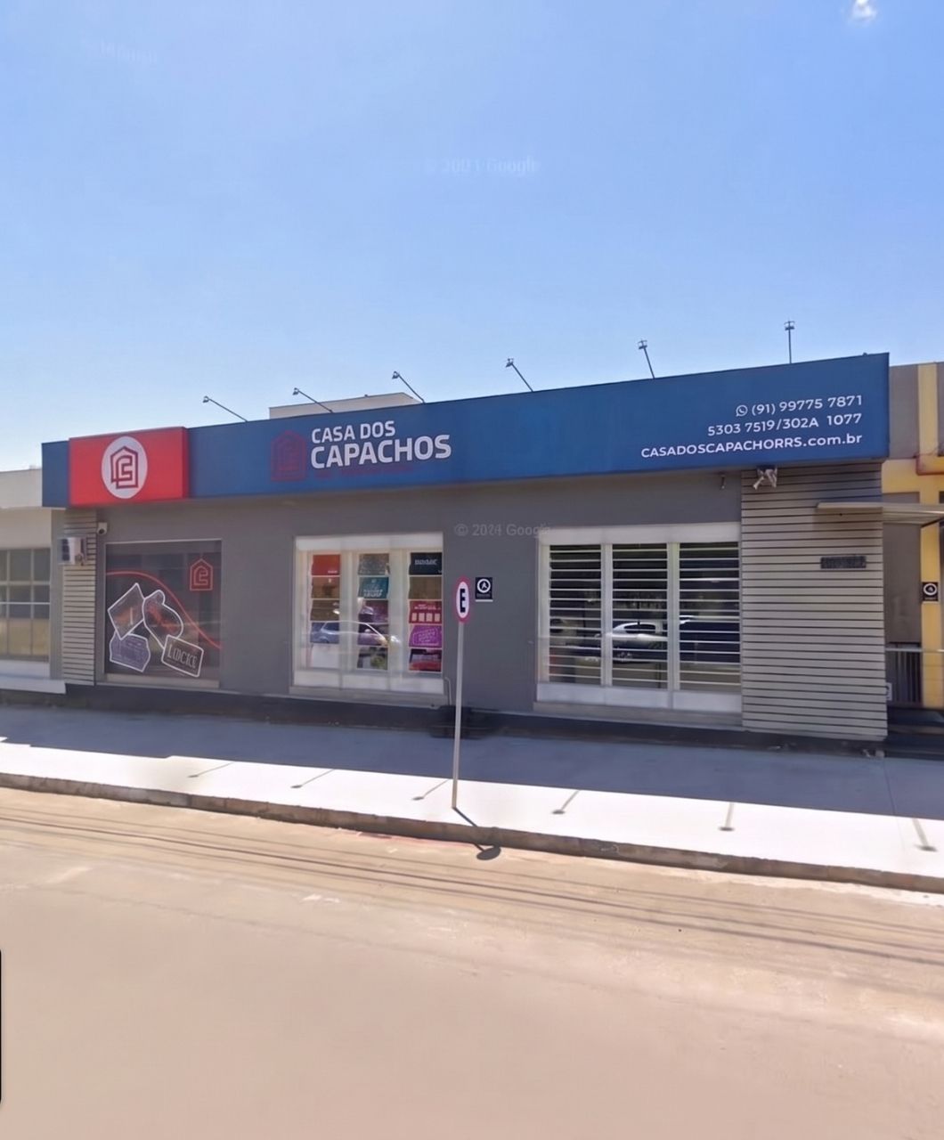 Fachada da loja Casa dos Capachos com letreiro azul e vermelho, logomarca vermelha e branca, entrada com vitrines de produtos, calçada em frente e céu azul claro, fotografia realista de rua comercial em dia ensolarado